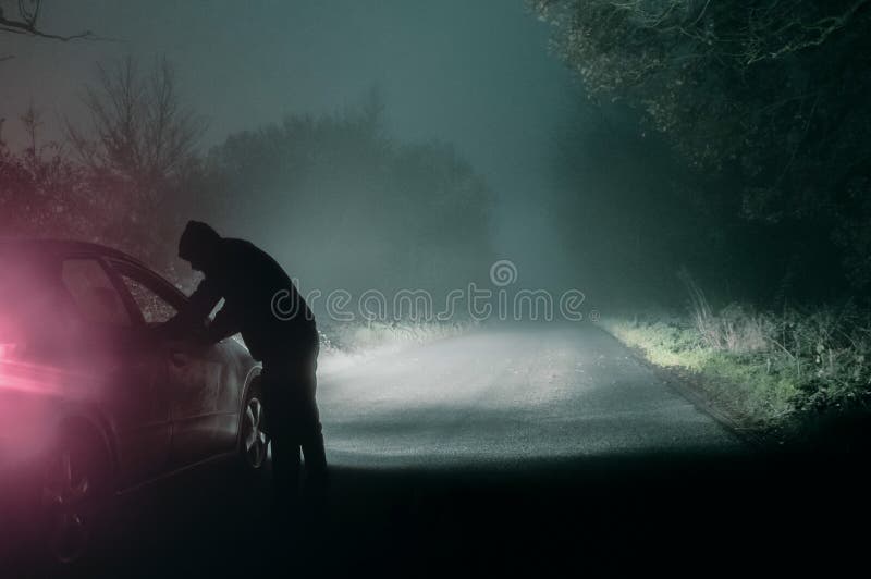 A lone hooded figure looking into a car. Silhouetted on a spooky, country foggy winters road at night. With a cold, grainy edit