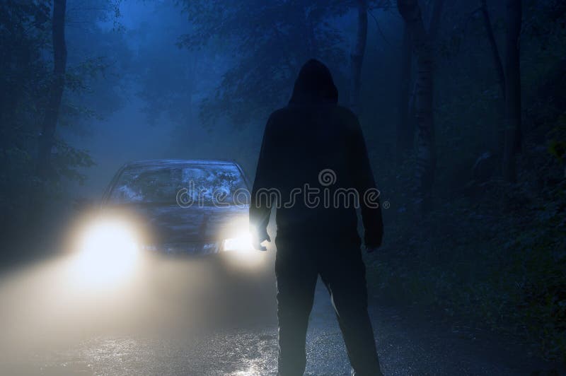 A lone hooded figure standing in front of car headlights in the middle of a country road on a spooky, eerie, moody foggy night.