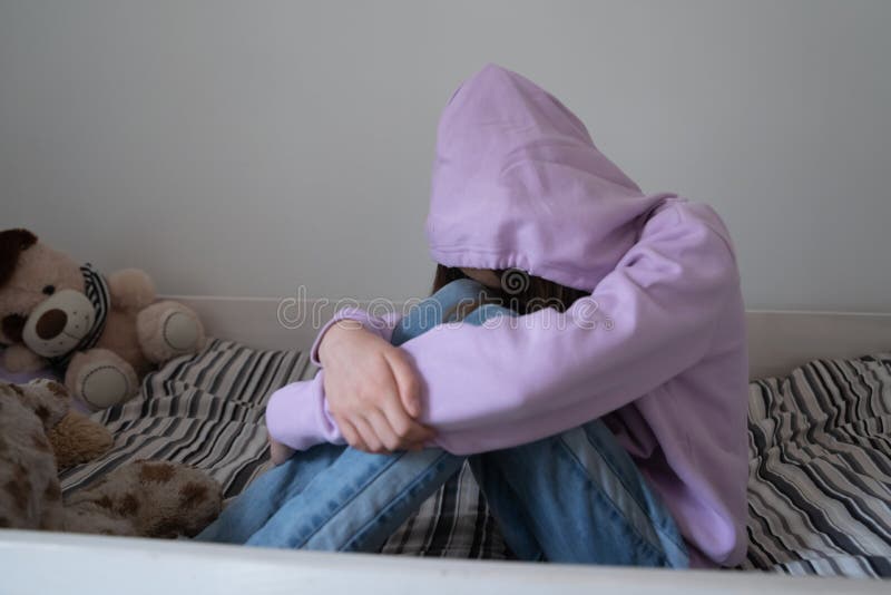 Lonely stressed teenage girl wearing hood sitting on bed alone, crying, thinking about troubles, hiding face, sad upset teenager feeling misunderstood and lonely, child and psychological problem. Lonely stressed teenage girl wearing hood sitting on bed alone, crying, thinking about troubles, hiding face, sad upset teenager feeling misunderstood and lonely, child and psychological problem