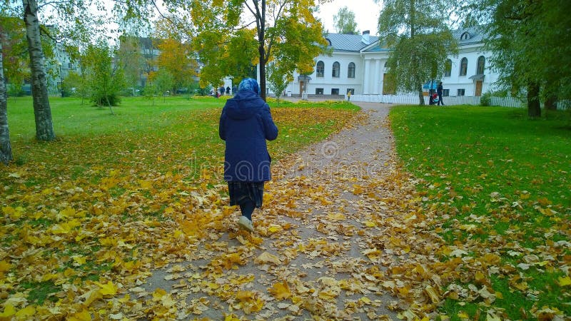 Lonely elderly woman walks in autumn park along path covered with fallen yellow orange maple leaves. Thoughts of old age, wilting