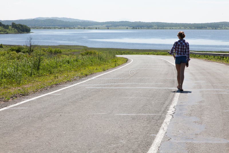 Lonely girl walking along the middle of the road on the background of the sea and the rural landscape.