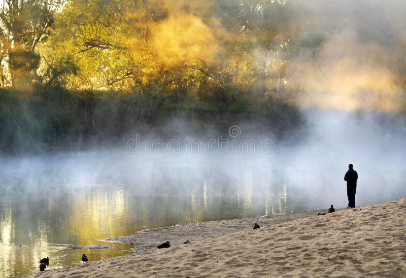 Un uomo in piedi da solo in pausa di alba, così come il sole è appena iniziando a salire tra gli alberi e riflettere attraverso la nebbia e la foschia l'acqua immobile di misty river.