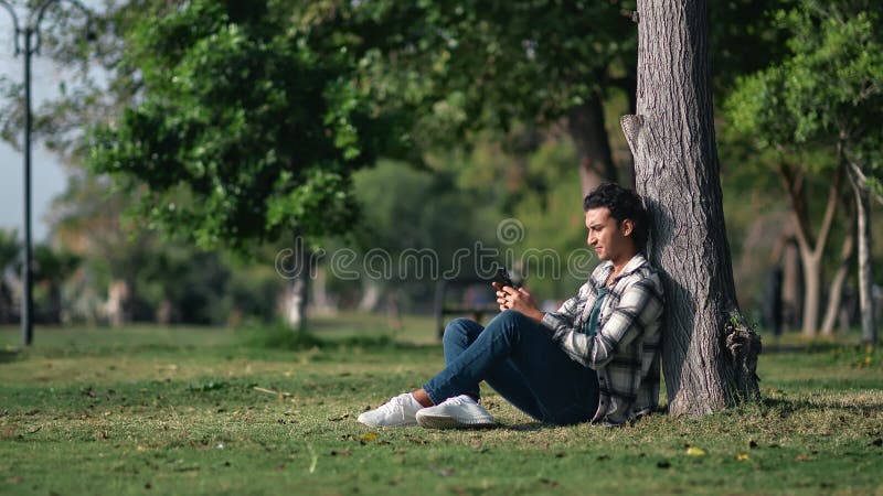 Lonely mixed race man use smartphone resting at summer sunny city park with green grass and trees stock video