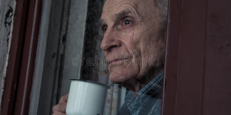 Lonely old retired man with white mug of coffeein hands looking through window