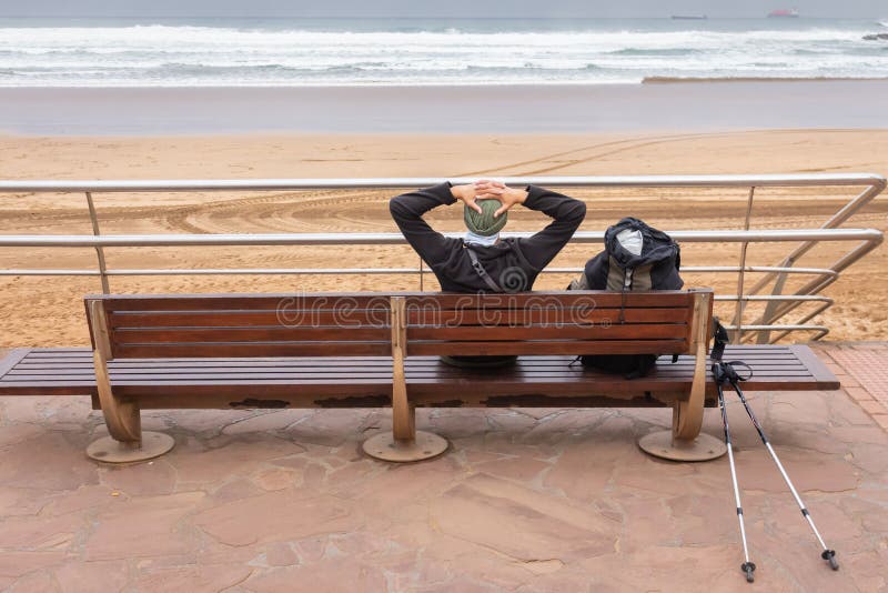 Lonely tourist have a rest on wooden bench on the seaside. Tired pilgrim with backpack and sticks on the beach. Pause and relax.