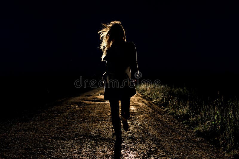 Blonde lonely young frightened woman on an empty night road runs away in the light of the headlights of her car