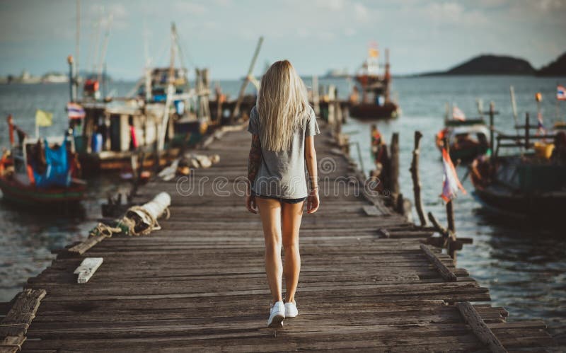 With long blonde hair coming through the old wooden pier in the rays of the setting sun. Back to the camera. Vacation in