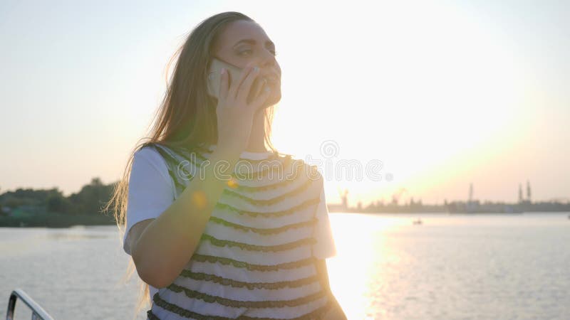 Long-haired girl talking on mobile phone on river embankment in sundown