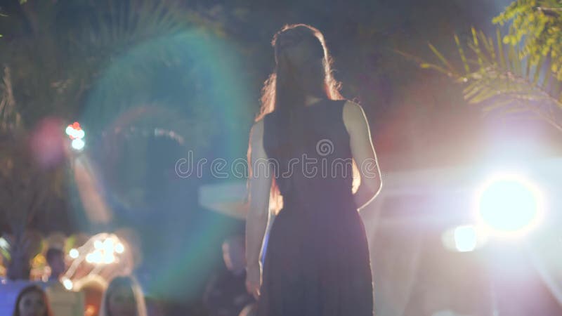 Long-haired woman in black dress at night outdoors in bright light, rear view