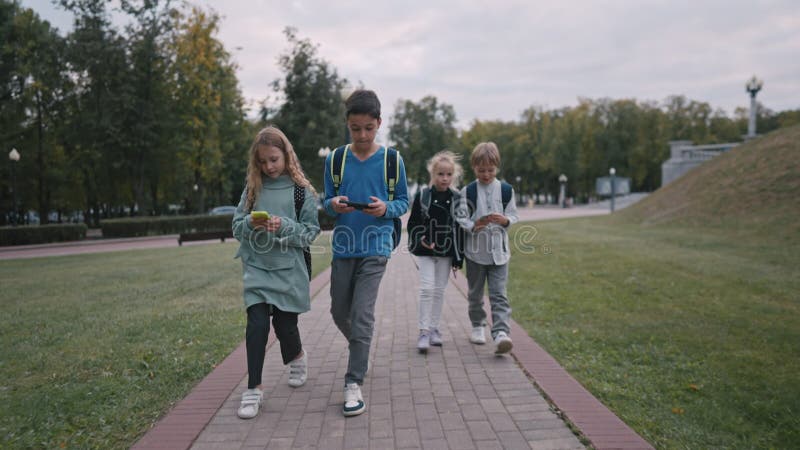 Long shot of four pupils with backpacks going out of school and talking. children use smartphones while walking from