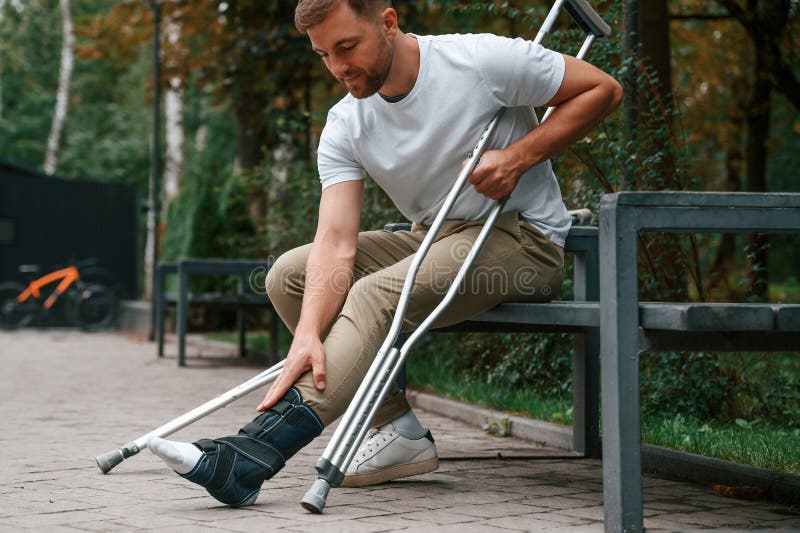 Looking at the Broken Leg. Man with Crutches is in the Park Outdoors