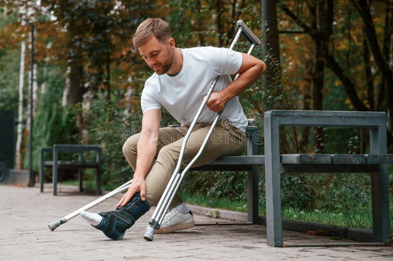 Looking at the Broken Leg. Man with Crutches is in the Park Outdoors