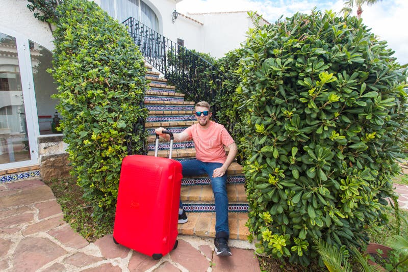Lopp-, turism- och feriebegrepp - lycklig man som sitter på trappa i soliga exponeringsglas som rymmer den röda resväskan royaltyfri bild Lopp-, turism- och feriebegrepp - lycklig man som sitter på trappa i soliga exponeringsglas som rymmer den röda resväskan royaltyfri bild
