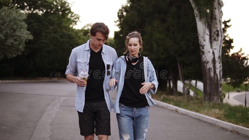 Los amigos jovenes hermosos en camisetas negras y camisas azules están caminando por el parque en la oscuridad de la tarde El hab