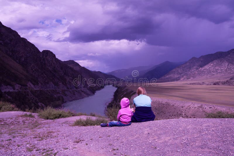 Little girl in a lilac jacket and girl-teenager in a violet jacket sit in mountains in a relaxation under the cloudy violet sky. Little girl in a lilac jacket and girl-teenager in a violet jacket sit in mountains in a relaxation under the cloudy violet sky