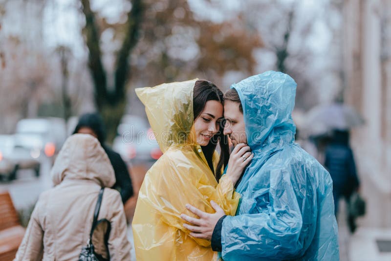 Los pares cari?osos rom?nticos, el individuo y su novia en los impermeables se colocan cara a cara en la calle bajo la lluvia imagenes de archivo Los pares cari?osos rom?nticos, el individuo y su novia en los impermeables se colocan cara a cara en la calle bajo la lluvia imagenes de archivo