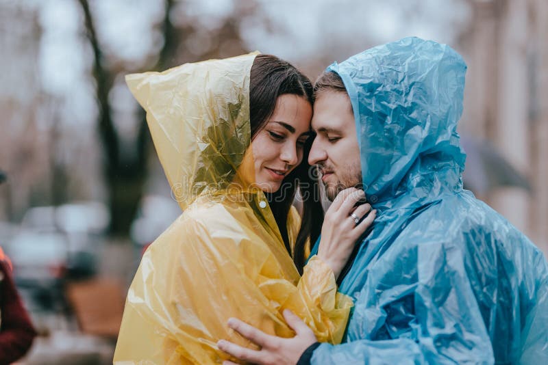 Los pares cari?osos rom?nticos, el individuo y su novia en los impermeables se colocan cara a cara en la calle bajo la lluvia imagen de archivo Los pares cari?osos rom?nticos, el individuo y su novia en los impermeables se colocan cara a cara en la calle bajo la lluvia imagen de archivo