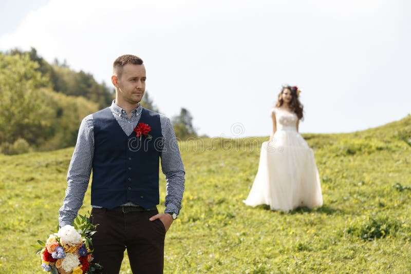 Los pares en la boda attire con un ramo de flores y el verdor está en las manos contra el contexto del campo en fotos de archivo libres de regalías