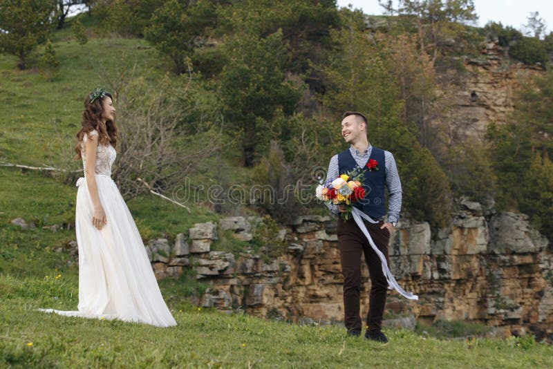 Los pares en la boda attire con un ramo de flores y el verdor está en las manos contra el contexto del campo en fotos de archivo