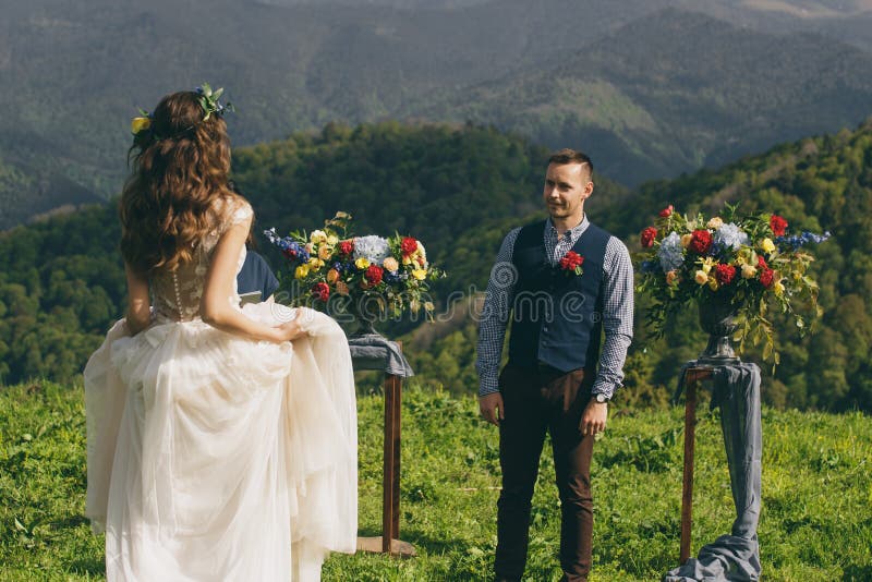 Los pares en la boda attire con un ramo de flores y el verdor está en las manos contra el contexto del campo en la puesta del sol, la novia