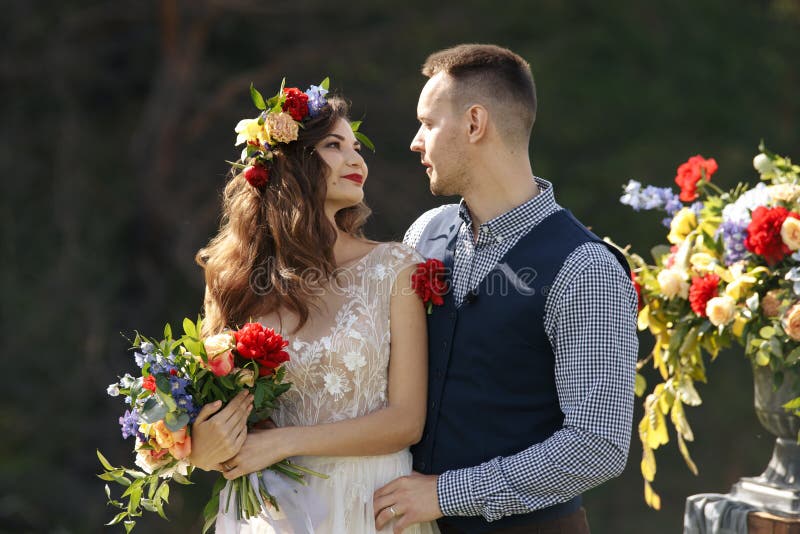 Los pares en la boda attire con un ramo de flores y el verdor está en las manos contra el contexto del campo en fotografía de archivo