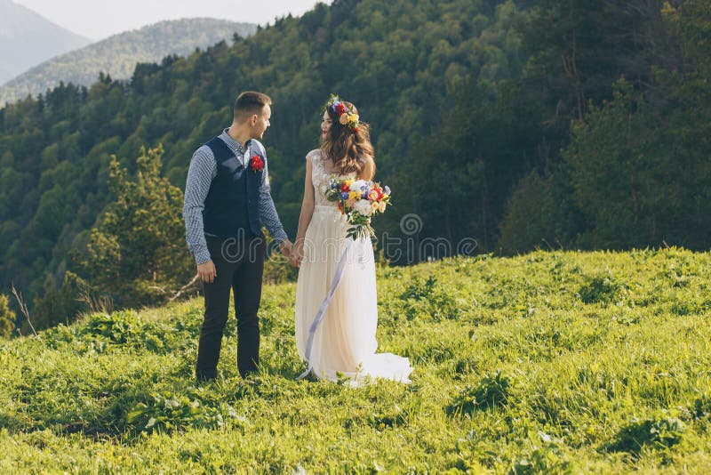 Los pares en la boda attire con un ramo de flores y el verdor está en las manos contra el contexto del campo en imagen de archivo libre de regalías