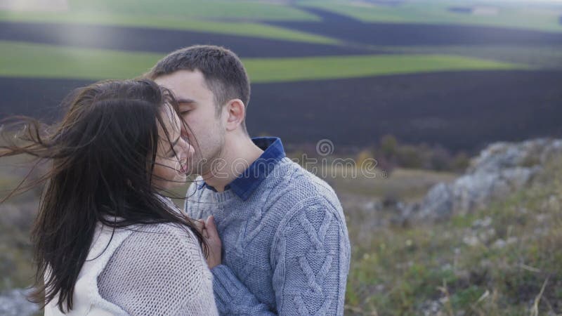 Los pares felices se colocan en la roca, los tactos y los besos en día ventoso 4K