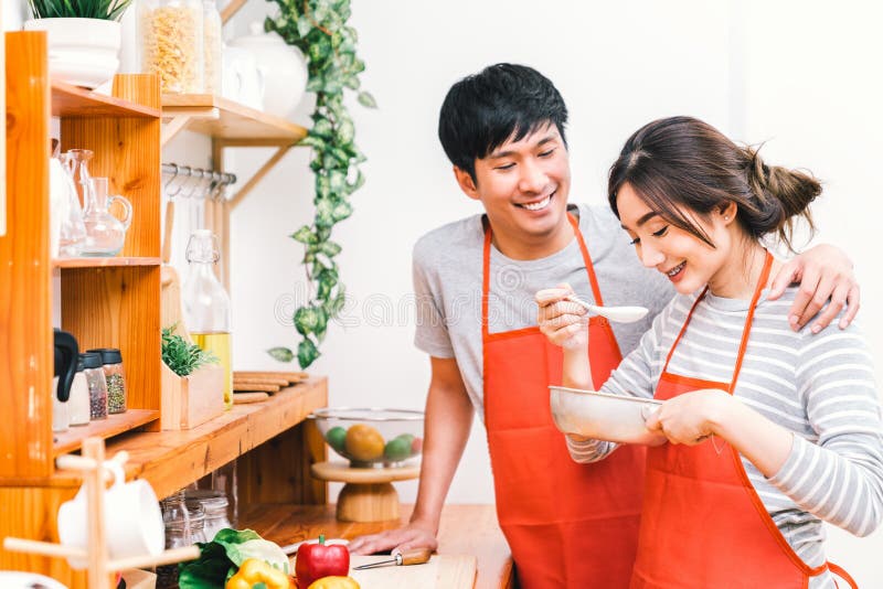 Los pares preciosos asiáticos jovenes que cocinan juntos en casa la cocina, llevan el delantal rojo que hace la comida del almuer fotografía de archivo