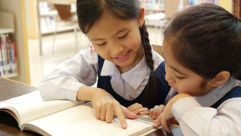 Los pequeños estudiantes asiáticos con el libro de lectura uniforme en biblioteca juntos, inclinan encima de cámara