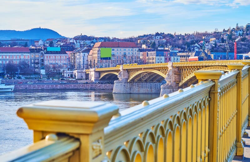 The closeup of the bright yellow metal rails of the Margaret Bridge against the Buda district and Danube River, Budapest, Hungary. The closeup of the bright yellow metal rails of the Margaret Bridge against the Buda district and Danube River, Budapest, Hungary