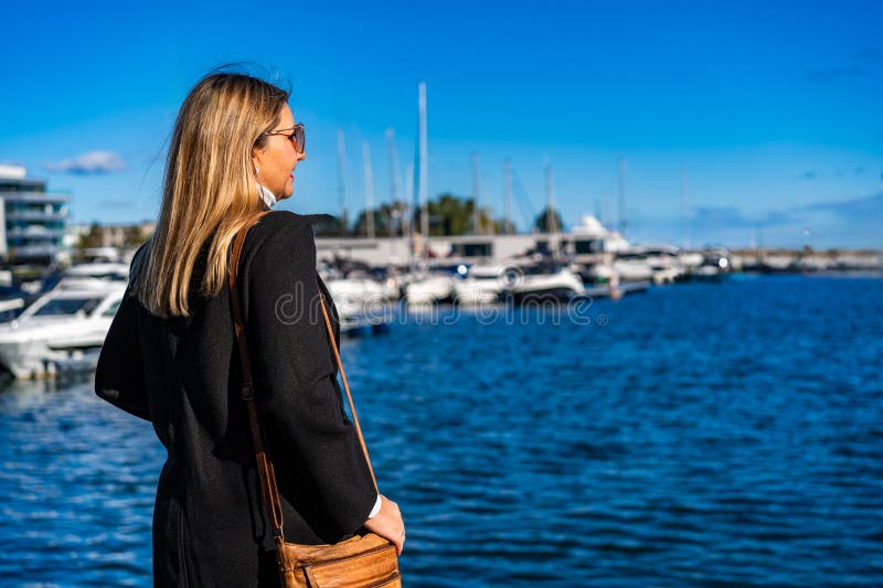 Loura-de-cabra-longa-de-cabelos-médios lindas usando bolsa de couro preto e óculos de sol em pé e olhando fotografia de stock