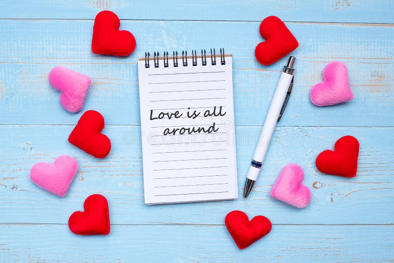 LOVE IS ALL AROUND word on note book and pen with red and pink heart shape decoration on blue wooden table background. Love, Wedding, Romantic and Valentineâ€™ s day concept