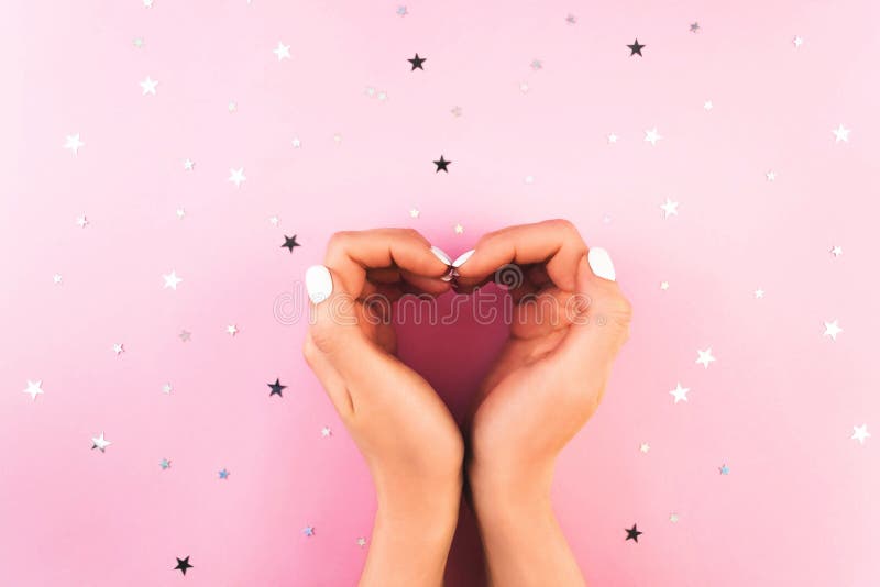 Love heart shape gesture. Two young woman`s hands with perfect white manicure on pink background with star confetti. Beauty, fashion, spa salon flat lay concept. Trendy wellness pastel nail gel polish. Love heart shape gesture. Two young woman`s hands with perfect white manicure on pink background with star confetti. Beauty, fashion, spa salon flat lay concept. Trendy wellness pastel nail gel polish