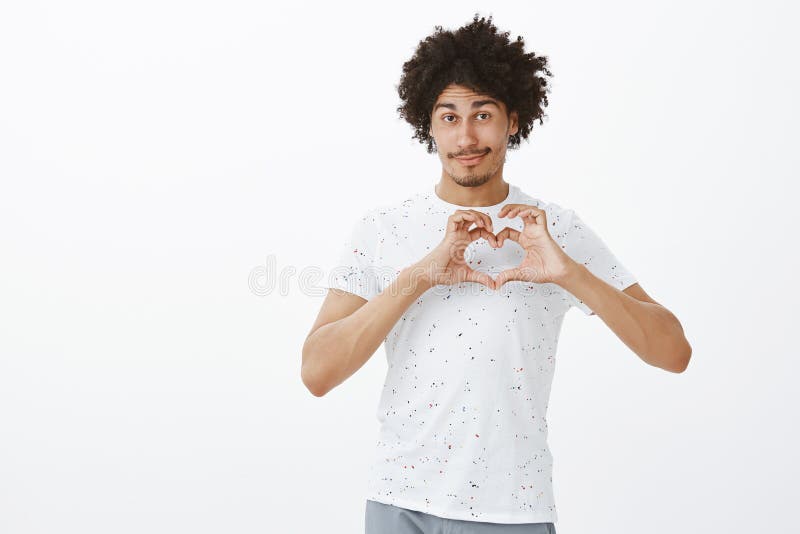 Love my friends. Cute friendly-looking relaxed hispanic male with tanned skin and afro hairstyle showing heart sign over royalty free stock photography Love my friends. Cute friendly-looking relaxed hispanic male with tanned skin and afro hairstyle showing heart sign over royalty free stock photography