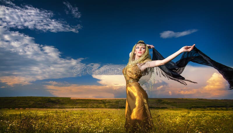 Lovely young lady posing dramatically with long black veil on green field. Blonde woman with cloudy sky in background - outdoor