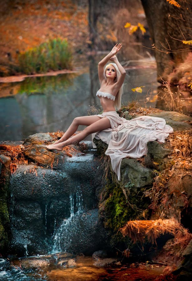 Lovely young lady sitting near river in enchanted woods. Sensual blonde with white clothes posing provocatively in autumnal park.