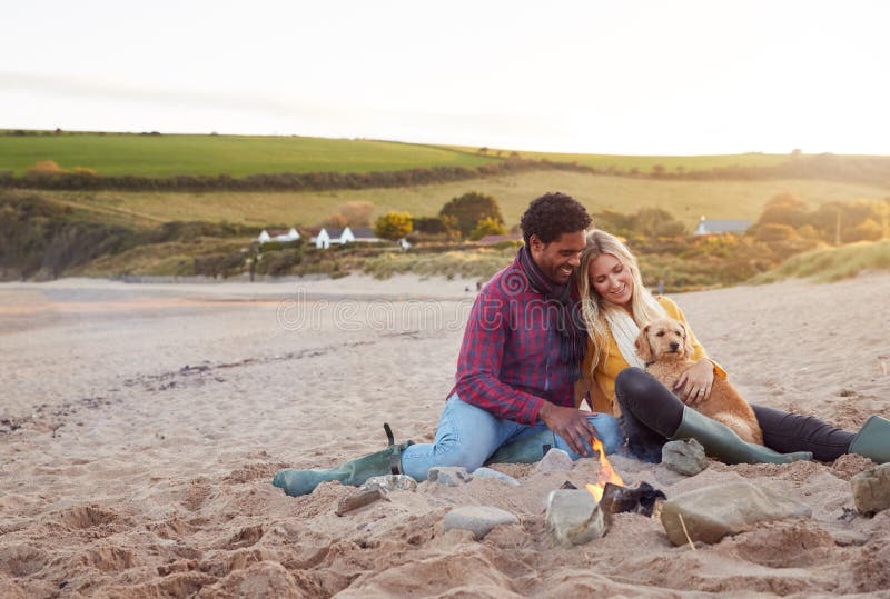 Loving Couple With Dog Relaxing By Fire On Winter Beach Vacation stock photos