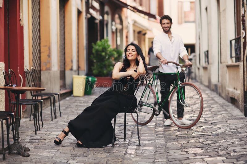 A loving couple in the old city. A woman in a black dress sits on a chair. The man behind her stands with a green bike royalty free stock photos A loving couple in the old city. A woman in a black dress sits on a chair. The man behind her stands with a green bike royalty free stock photos