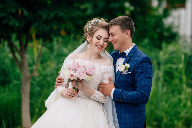 A loving couple walks in a green park on their wedding day. The newlyweds embrace and laugh, gently touch each other`s