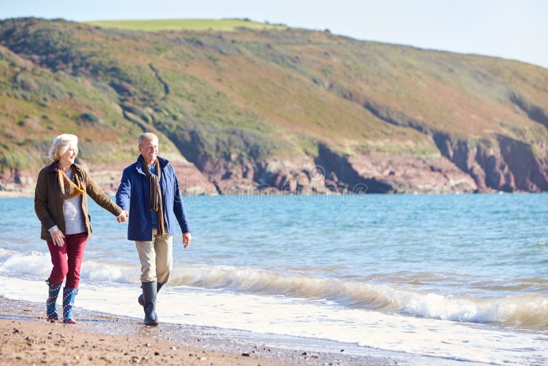 Loving Senior Couple Holding Hands As They Walk Along Shoreline Of Beach By Waves