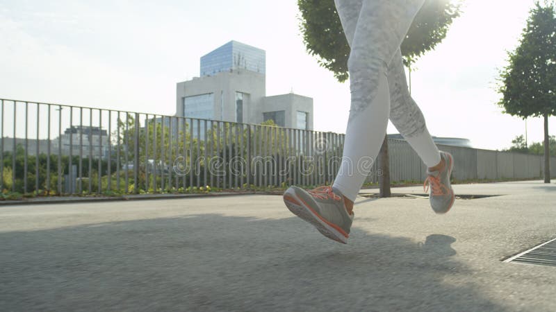 LOW ANGLE, CLOSE UP: Fit girl in tights runs down green avenue in modern city. stock photography