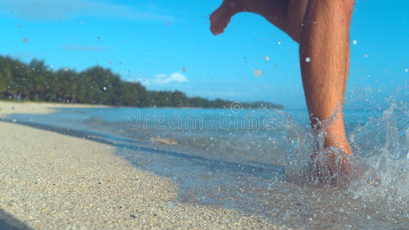 LOW ANGLE: Glassy drops of sea water fly around unrecognizable man`s feet as he runs down the sunny beach. stock photos