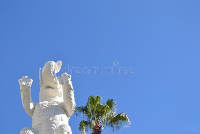 Low Angle Shot Of A White Elephant Statue Near A Palm Tree Under A Clear Blue Sky