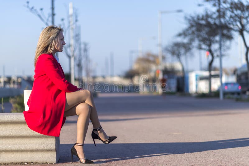 Low angle view of a elegant blonde woman in red jacket sitting on a bench outdoors while looking away in sunny day