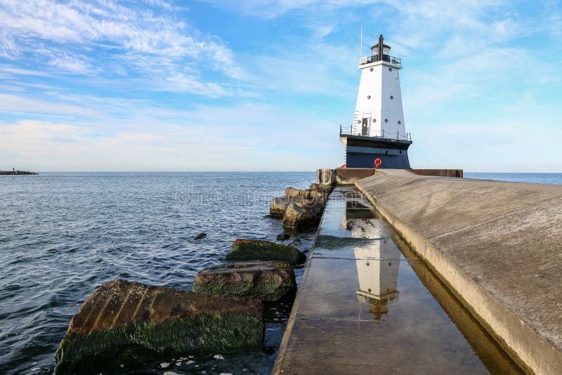 North Breakwater Pier light in Ludington Michigan is reflected in standing water on the pier walkway. Moss clings to rocks in the foreground and clouds swirl through the blue sky above. North Breakwater Pier light in Ludington Michigan is reflected in standing water on the pier walkway. Moss clings to rocks in the foreground and clouds swirl through the blue sky above.