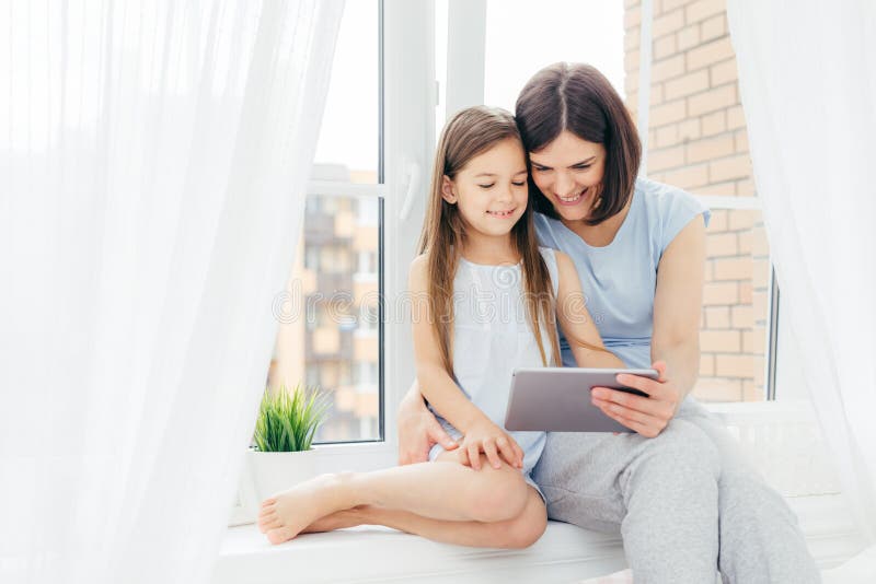 People, technology, family, children concept. Positive young other and her small daughter sit on window sill, hold digital tablet, watch interesting cartoon, pose near window with white curtains. People, technology, family, children concept. Positive young other and her small daughter sit on window sill, hold digital tablet, watch interesting cartoon, pose near window with white curtains