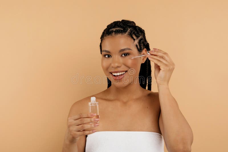 Lugn tusal afrikansk amerikansk dam som applicerar serum på ansiktet isolerat på beige studions bakgrund. livsstil royaltyfria bilder