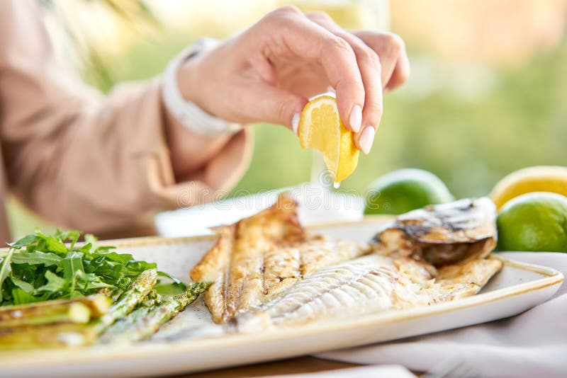 Lunch in a restaurant, a woman eats roasted dorado fish with grilled asparagus. Dish decorated with a slice of lemon