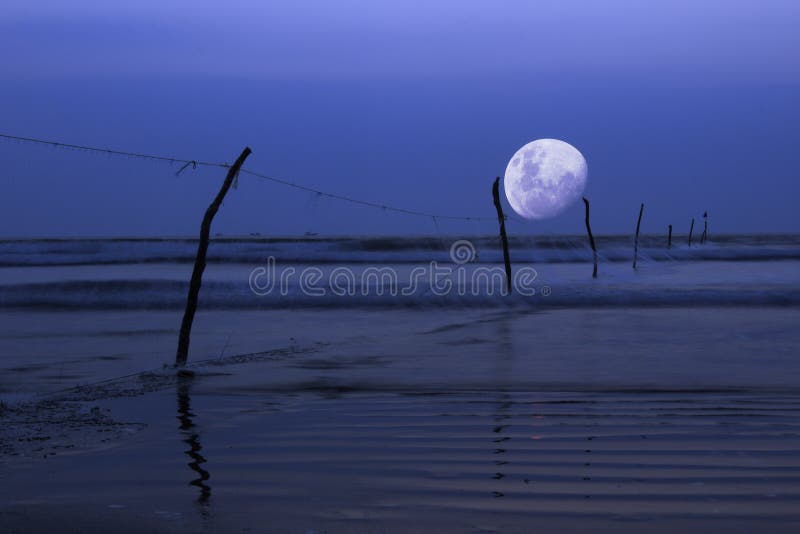 A blue moon over blue night scene and a slow dreamy ocean with fishing net poles in the ocean going deep into the ocean, half moon rising or setting over the water. A blue moon over blue night scene and a slow dreamy ocean with fishing net poles in the ocean going deep into the ocean, half moon rising or setting over the water.