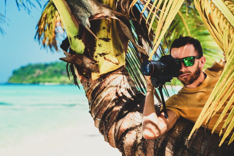 Lustige männliche Paparazziphotographfelle hinter einem Baum auf einem tropischen Strand, zum von Fotos einer versteckten Kamera lizenzfreie stockfotografie Lustige männliche Paparazziphotographfelle hinter einem Baum auf einem tropischen Strand, zum von Fotos einer versteckten Kamera lizenzfreie stockfotografie